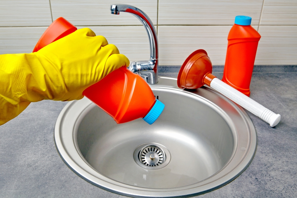 Hands in yellow rubber gloves hold a plunger and a pipe cleaner against the background of a kitchen sink.